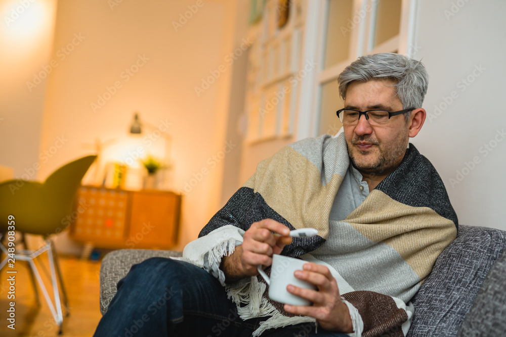 man sitting sofa holding cup of tea and looking at thermometer man sitting sofa holding cup of tea and looking at thermometer