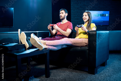 Young couple playing video games with gaming console sitting on the couch in the dark room of the playing club