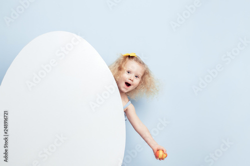 Funny little curly girl in the light-blue dress with bunny ears on her head looks out from behind a big white egg against a blue wall. Easter bunny