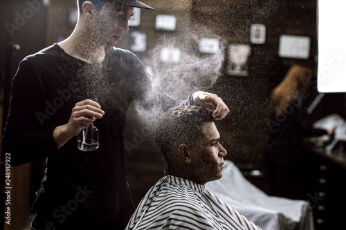 The barber dressed in a black clothes is doing the hair styling to the stylish black-haired  man sitting in the armchair in a barbershop
