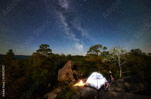 Woman hiker resting on rocky mountain top beside camp  bonfire and tourist tent at summer night  enjoying view of sky full of stars and Milky way. On background starry sky  big boulders and trees