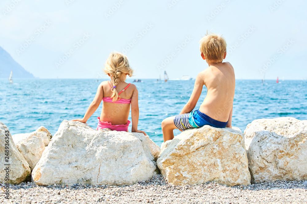 Children girl and boy (siblings) sitting on the rock and enjoying the view of sea at summer holiday. Concept of summer family vacation