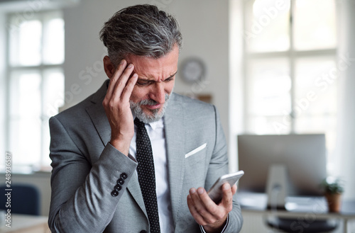 A frustrated businessman with smartphone standing in an office, reading bad news. photo
