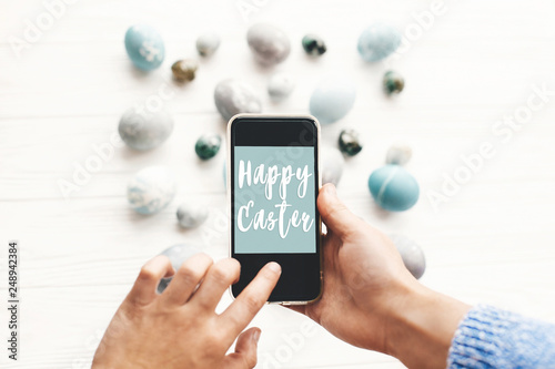 Photography Happy Easter text sign on phone screen in hands above stylish easter eggs on rustic white wooden table
