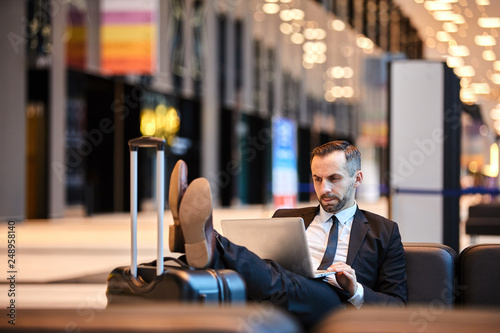 Young serious businessman in suit browsing in the net while resting on sofa in airport lounge