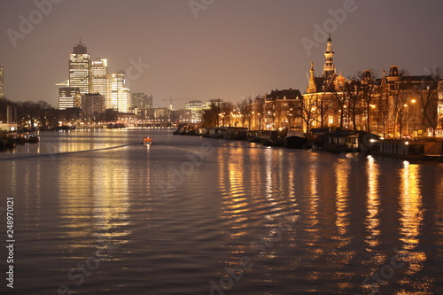 AMSTERDAM, NETHERLANDS - NOVEMBER 21, 2018: Illuminated buildings reflected in calm water, amazing cityscape with boat on Amstel canal at night © Edwin Butter