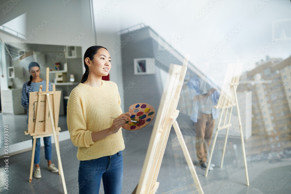 Pretty young creative girl in casualwear looking at painting on easel while working individually in studio