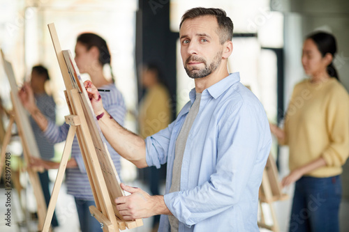 Young man in shirt looking at you while standing by easel and learning to paint and make sketches