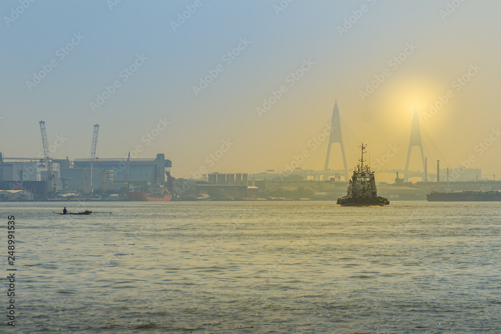 Fototapeta premium Commercial ship sailing on the river near a bridge during sunset at the estuary of Chaophraya river, Samut Prakarn province, Thailand.