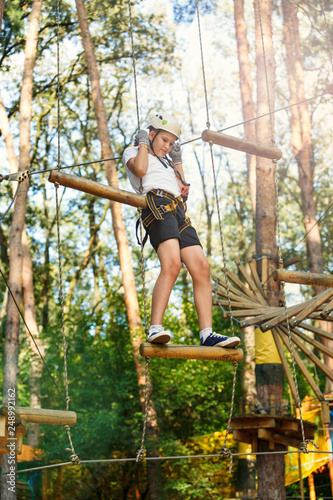Cheerful cute young boy in white t shirt and white helmet in adventure rope park at sunny summer day. Active lifestyle, sport, holidays for children