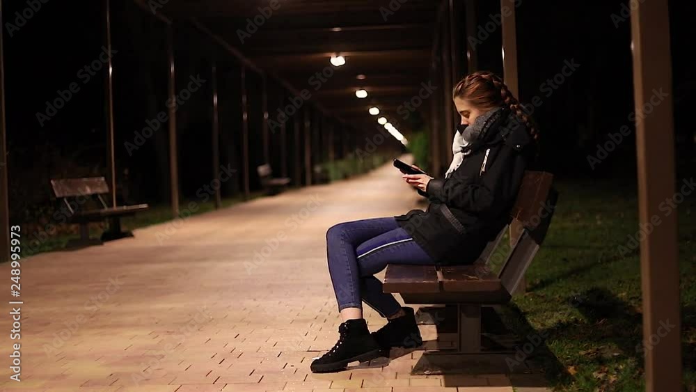 young woman alone looking their smart phone at night in a park bench slow motion young woman alone looking their smart phone at night in a park bench slow motion