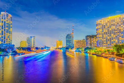 Beautiful night view during twilight with cruises and modern business buildings along the Chao Phraya river under blue sky background at Taksin Bridge, Bangkok, Thailand.