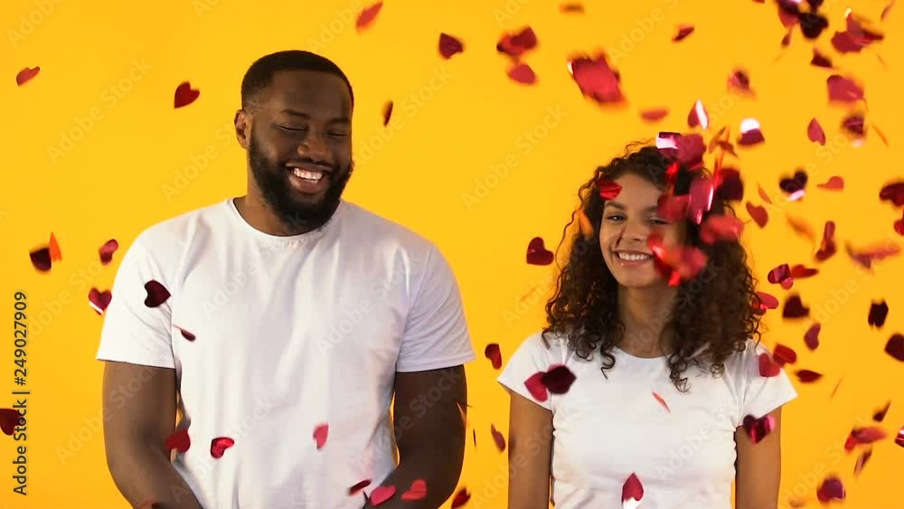 Cheerful Afro-American couple standing under falling heart-shaped confetti Cheerful Afro-American couple standing under falling heart-shaped confetti