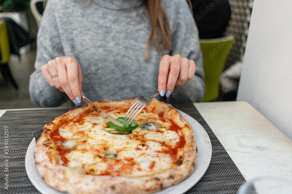 Woman eats with knife and fork a pizza Margherita with mozzarella tomatoes and basil. Neapolitan pizza from wood-burning stove. lunch in an Italian restaurant. Table near to a large window. Woman eats with knife and fork a pizza Margherita with mozzarella tomatoes and basil. Neapolitan pizza from wood-burning stove. lunch in an Italian restaurant. Table near to a large window.
