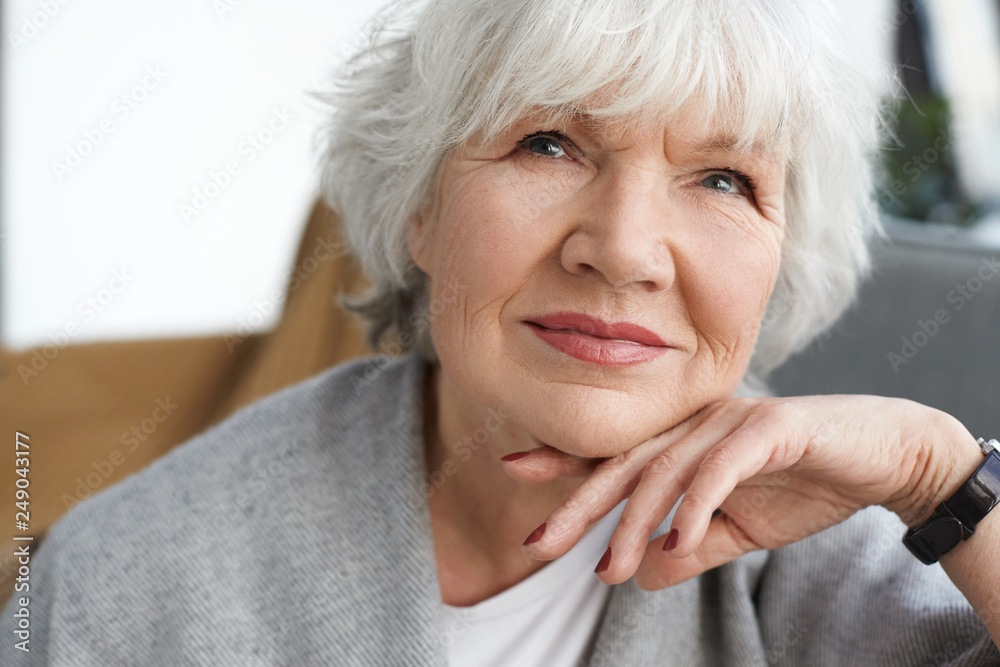 Close up portrait of beautiful positive middle aged Caucasian lady on retirement daydreaming at home, thinking about her grandchildren. Elegant gray haired grandmother spending time indoors Close up portrait of beautiful positive middle aged Caucasian lady on retirement daydreaming at home, thinking about her grandchildren. Elegant gray haired grandmother spending time indoors