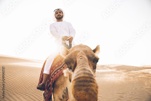 Man wearing traditional clothes, taking a camel out on the desert sand, in Dubai