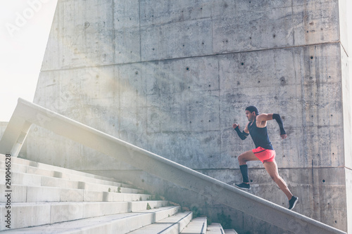 Muscular hispanic dark-skinned male athlete running up a flight of stairs with speed, sporty young man in black t-shirt training or working out outdoors while jogging up the steps. Sunlight, filtered.