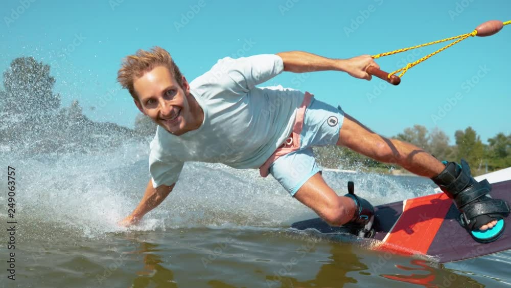 CLOSE UP, TIME REMAP: Handsome Caucasian man wakeboarding does a hand drag and splashes water while speeding past the camera. Smiling wakesurfer riding in the waterski cable park on sunny summer day.
