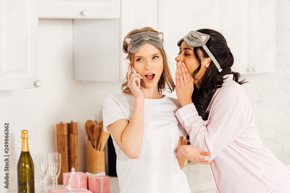 african american girl whispering secret to beautiful girl in sleeping mask talking on smartphone during pajama party african american girl whispering secret to beautiful girl in sleeping mask talking on smartphone during pajama party