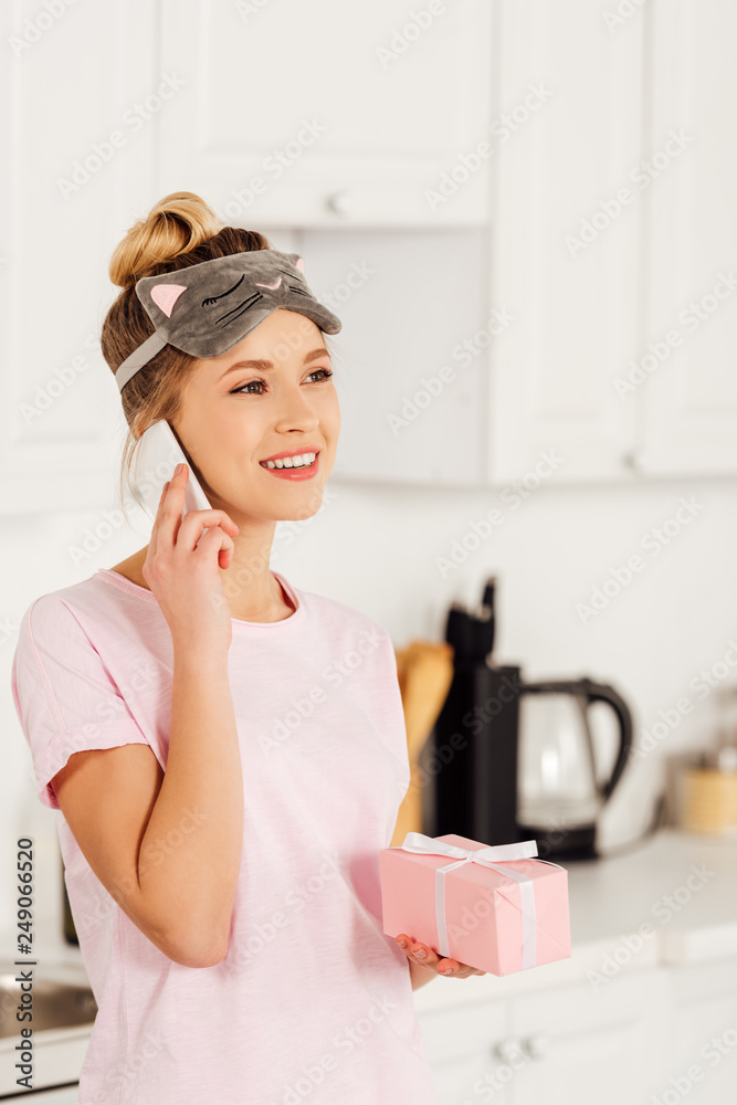 beautiful girl in pajamas and sleeping mask talking on smartphone while holding gift box at home in kitchen beautiful girl in pajamas and sleeping mask talking on smartphone while holding gift box at home in kitchen