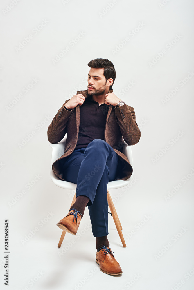 Confident in his style. Stylish dark-haired man sitting on a chair, crossing his legs and looking away isolated over white background Confident in his style. Stylish dark-haired man sitting on a chair, crossing his legs and looking away isolated over white background