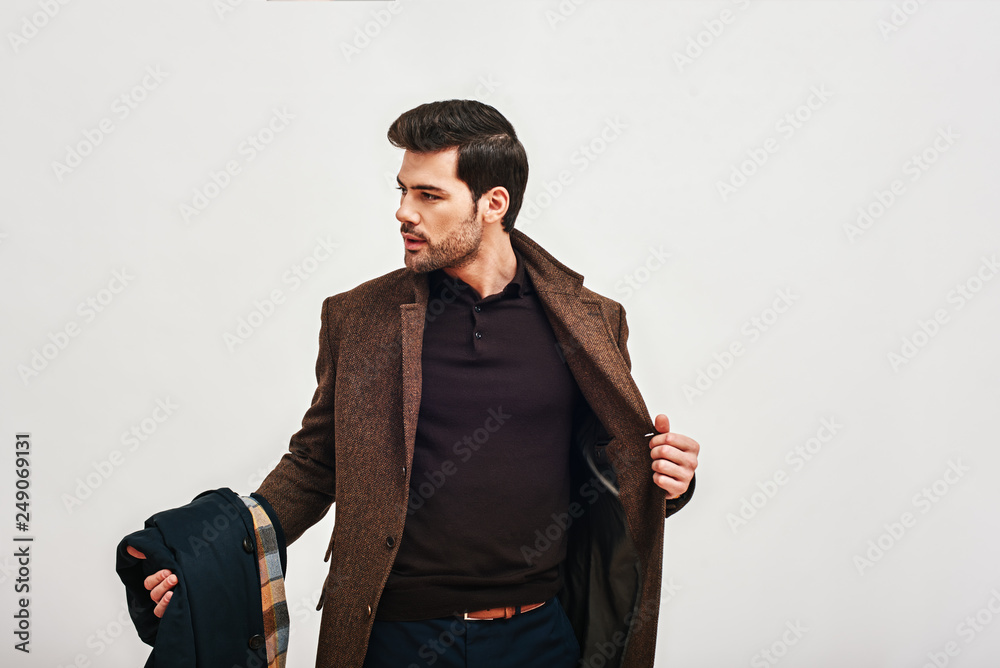 Always perfect. Stylish dark-haired man standing, holding his coat and looking away isolated over white background - obrazy, fototapety, plakaty 