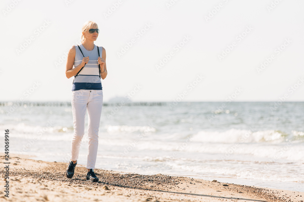 Young, beautiful blonde girl walking at the beach. Stylish woman in white pants and sunglasses near sea