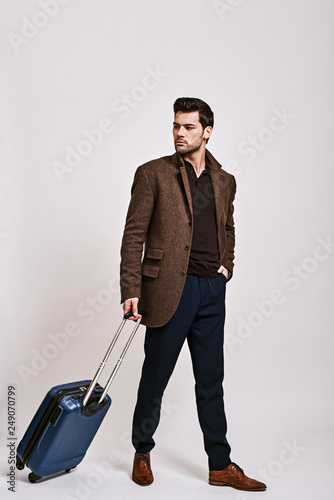 Ready to travel. Stylish dark-haired man standing with suitcase and looking away isolated over grey background