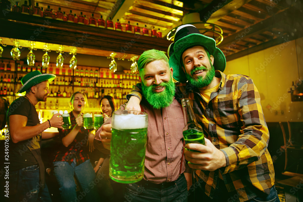 Obraz premium Saint Patrick's Day Party. Happy friends is celebrating and drinking green beer. Young men and women wearing a green hats. Pub Interior.