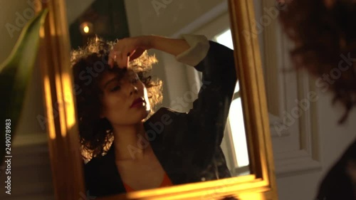 A beautiful woman with dark curly hair in a black jacket stands at the mirror and straightens her hair, touches her hair with her hands, close-up, portrait photography