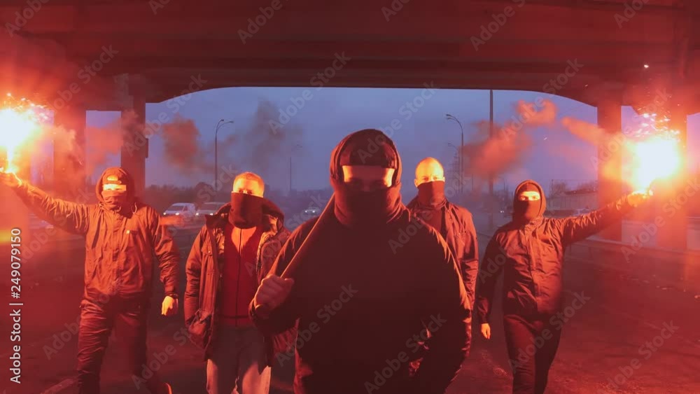 Group of young men in balaclavas with red burning signal flare walking on the road under the bridge, slow motion