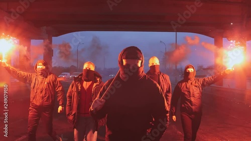 Group of young men in balaclavas with red burning signal flare walking on the road under the bridge, slow motion photo