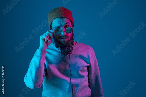 Enjoying his favorite music. Happy young stylish man in hat and sunglasses with headphones listening and smiling while standing against blue neon background