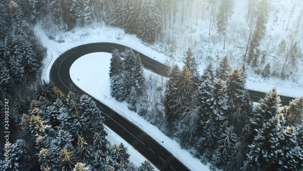 Overhead aerial top view over car travelling on hairpin bend turn road in mountain winter snow covered forest. White pine tree woods. Snowy street path establisher. 4k drone flight sidewards