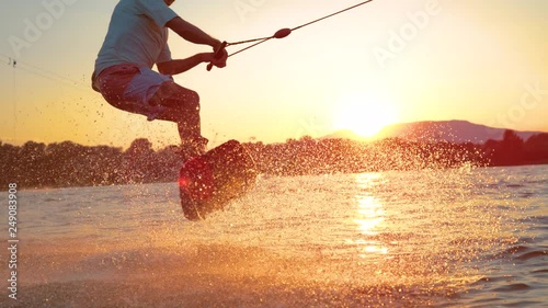 SLOW MOTION, SUN FLARE, CLOSE UP: Cool wakeboarder does 180 ollie while speeding across the lake at sunset. Athletic man on vacation having fun wakeboarding in the cool wake park on sunny morning.
