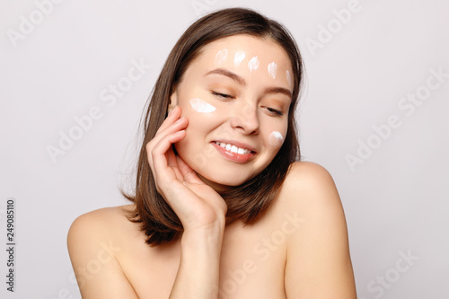 Close up beauty portrait of a laughing beautiful half naked woman applying face cream and looking away isolated over white background