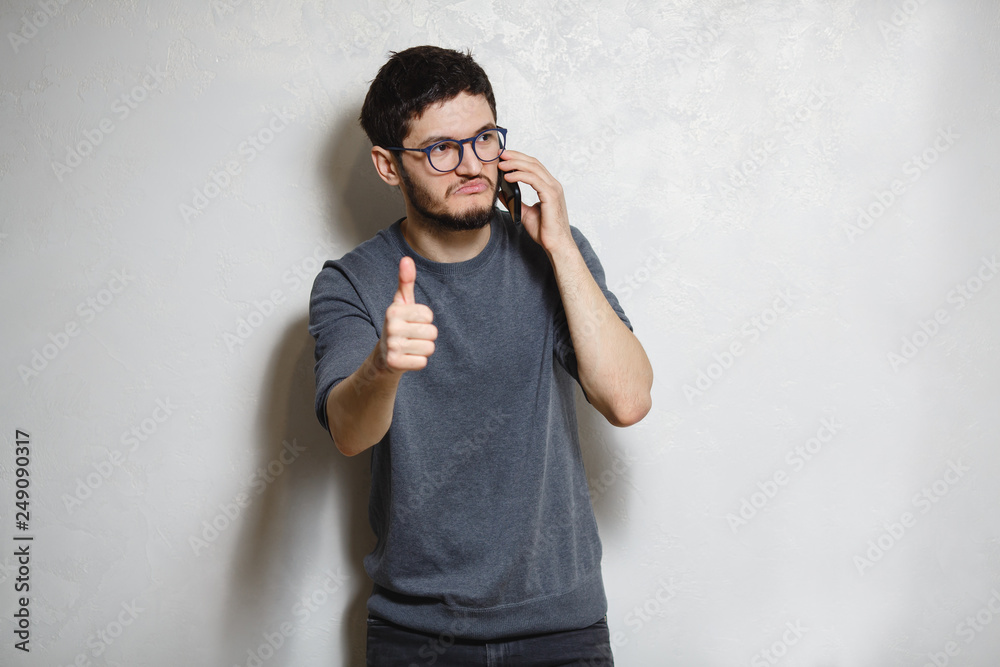 Portrait of young confident man with smartphone in hand showing thumb up, over white textured background. Portrait of young confident man with smartphone in hand showing thumb up, over white textured background.