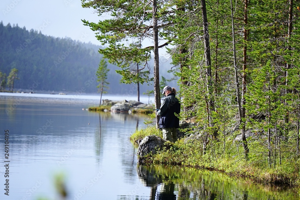 Man fishing, casting a fishing rod, in the lake in Lapland, Finland. Calm water, lake surrounded by trees.  