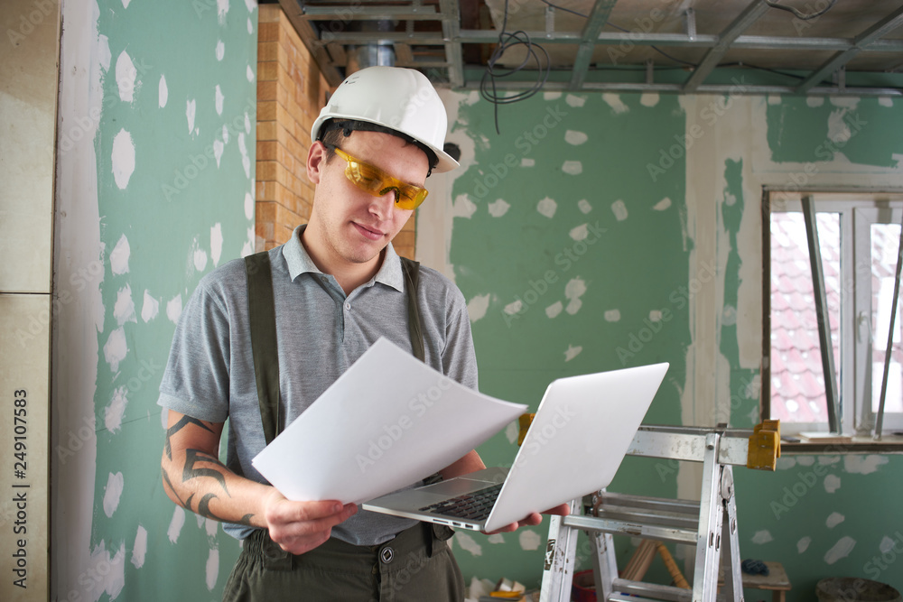 Room repair. Builder in helmet and glasses stands with a laptop and drawings in his hands on the background of the construction