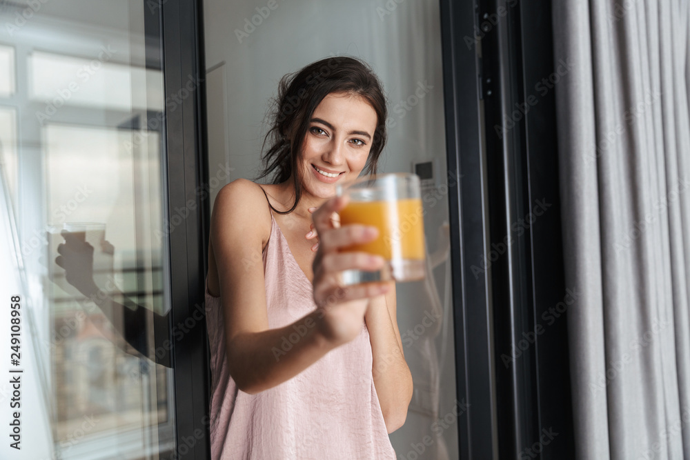 Photo of pretty woman 20s wearing silk leisure clothing, holding glass with orange juice in flat Photo of pretty woman 20s wearing silk leisure clothing, holding glass with orange juice in flat