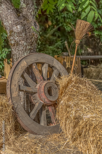 View of scenery in medieval fair, with traditional wagon wheel leaning against a tree and bales of straw with traditional broom