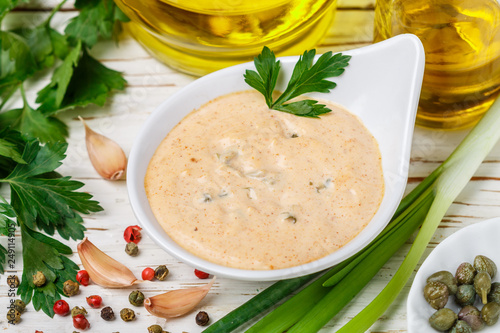 Homemade traditional French remoulade sauce in a White bowl with ingredients-capers, parsley, green onion, mustard, fragrant vinegar, olive oil, garlic on a light wooden table. dip. Selective focus photo