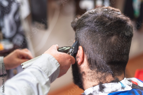 Handsome bearded man, getting haircut by barber, with electric trimmer at barbershop .