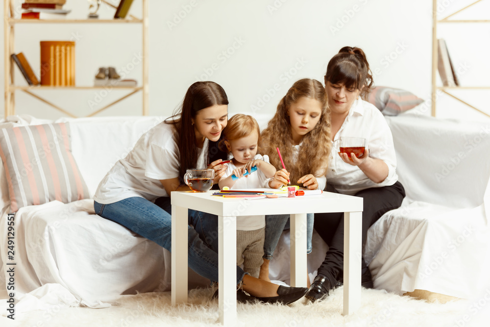 Two little girls their attractive young mother and their charming grandmother sitting on sofa and spending time together at home. Generation of women. International Women's Day. Happy Mother's Day. Two little girls their attractive young mother and their charming grandmother sitting on sofa and spending time together at home. Generation of women. International Women's Day. Happy Mother's Day.