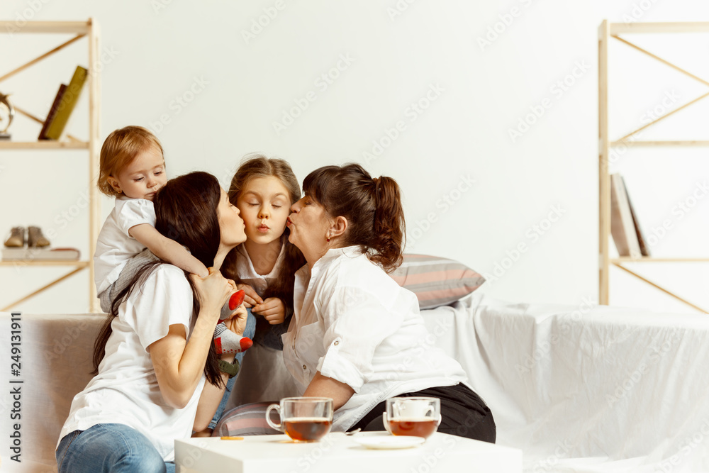 Two little girls their attractive young mother and their charming grandmother sitting on sofa and spending time together at home. Generation of women. International Women's Day. Happy Mother's Day. Two little girls their attractive young mother and their charming grandmother sitting on sofa and spending time together at home. Generation of women. International Women's Day. Happy Mother's Day.