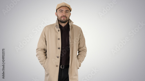 Confident man in trench holding hands in the pockets and walking on gradient background.