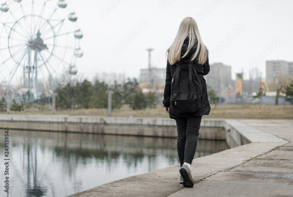 young girl with blond hair in black clothes with a backpack goes along the river bank to an amusement park