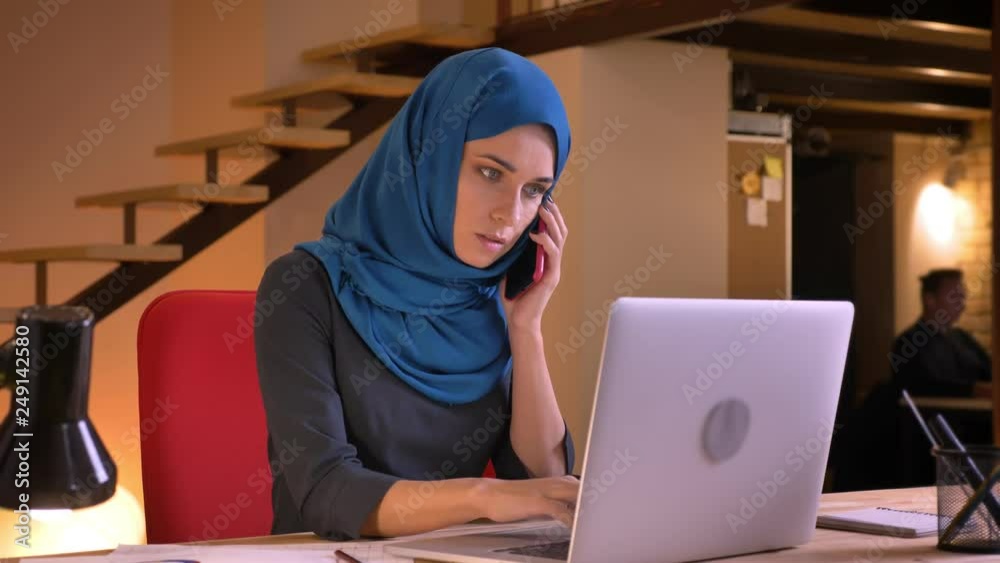 Closeup portrait of young attractive arabian female employee having a formal conversation on the phone while typing on the laptop Closeup portrait of young attractive arabian female employee having a formal conversation on the phone while typing on the laptop