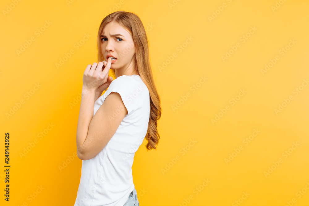 The excited girl is frightened by something, bites her nails, has a restless facial expression, stands on a yellow background, The excited girl is frightened by something, bites her nails, has a restless facial expression, stands on a yellow background,