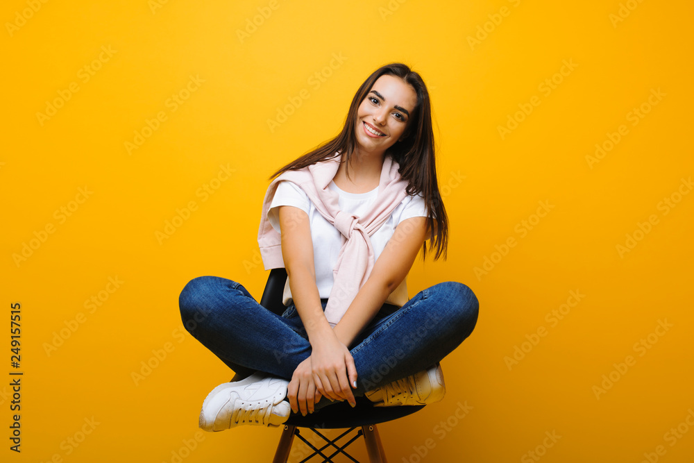 Amazing caucasian woman sitting on a chair dressed in casual clothes and smiling while leaning her head away with crossed hands in front o yellow background.